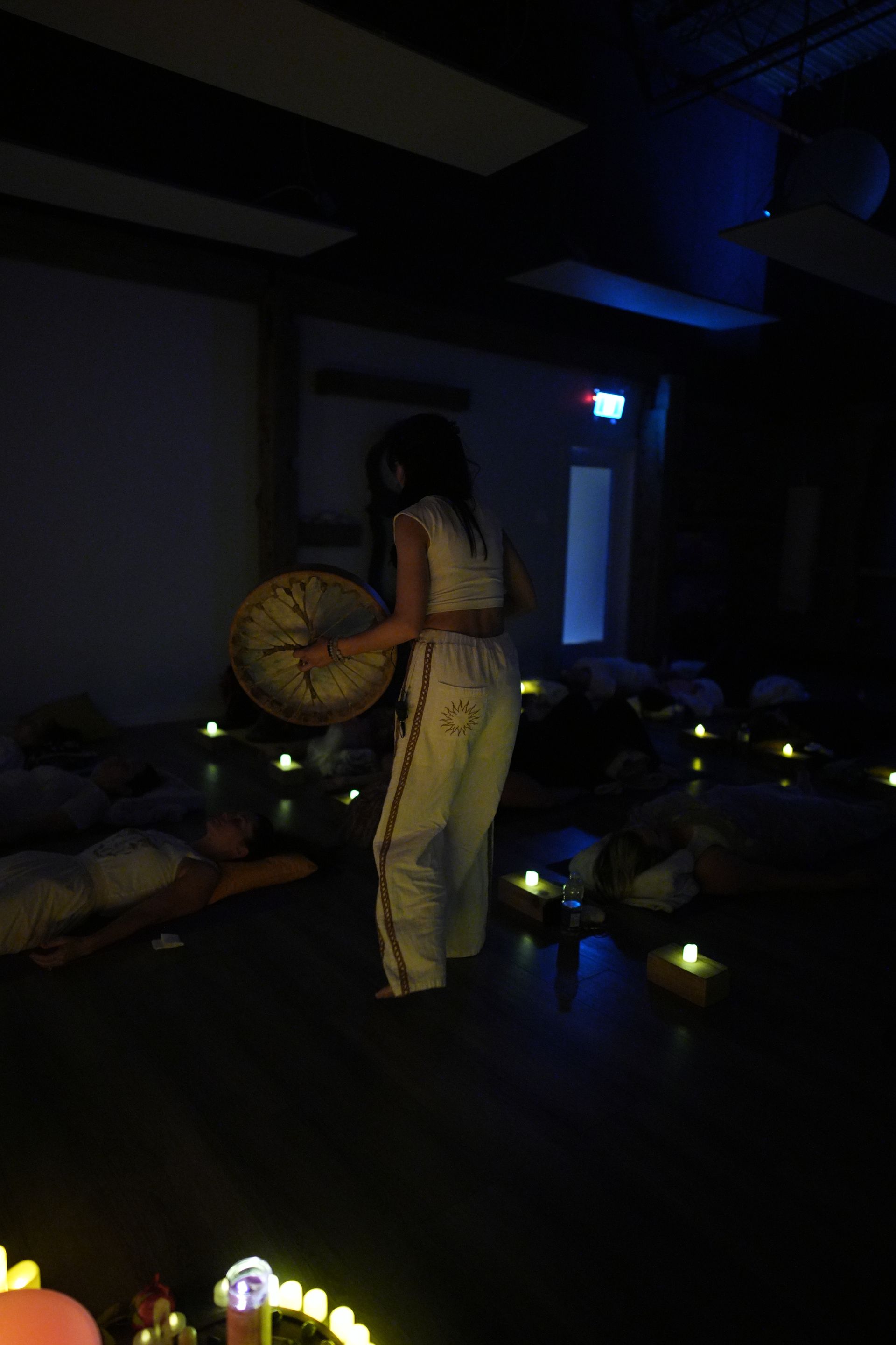 Woman playing drum in dimly lit room. People lie on the floor, surrounded by candles.