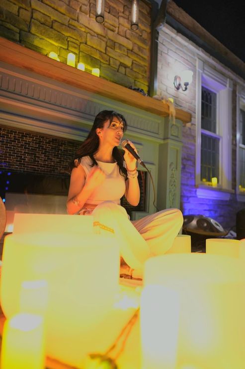 Woman singing into a microphone, seated, surrounded by lit candles, exterior, nighttime.