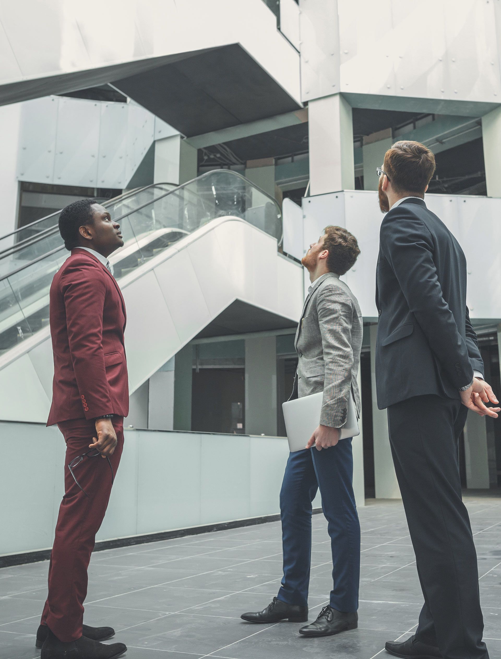 Three men in suits are standing in front of a building looking up.