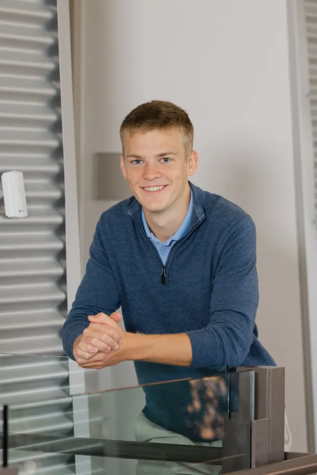 Young person in blue sweater, leaning on a glass surface, smiling in a modern setting.