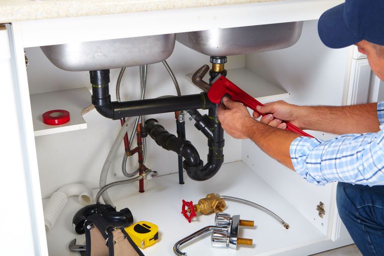 Plumber tightening under-sink pipes with a red wrench in a white cabinet closet