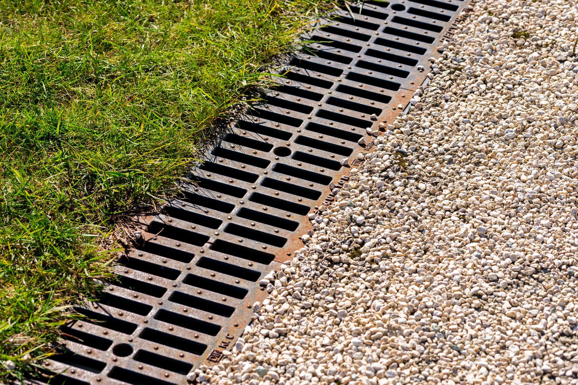 Black drainage grate between green grass and gravel along a sloped edge