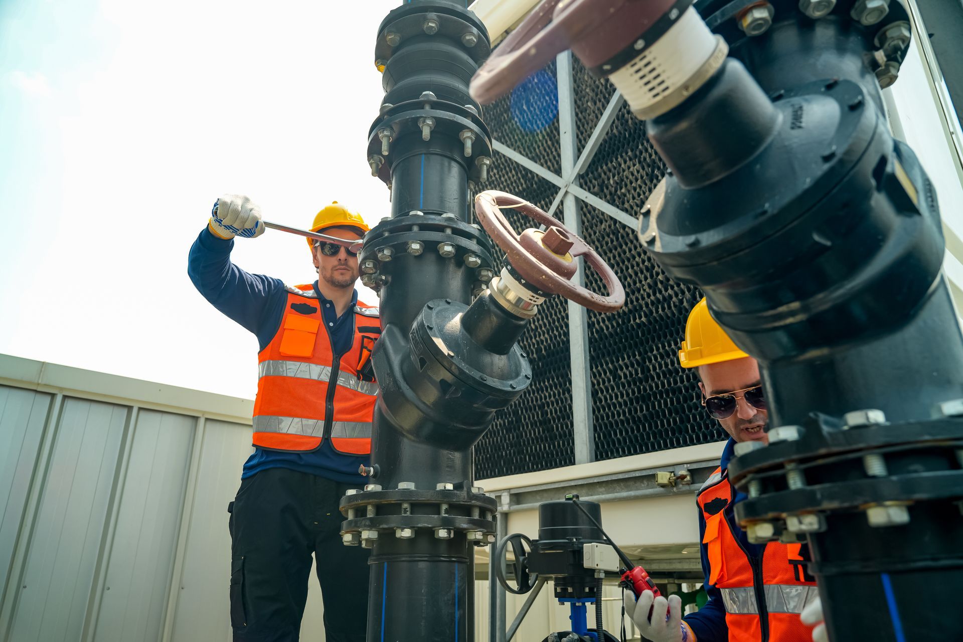 Two utility workers in orange safety vests inspecting large industrial pipes and valves outdoors.