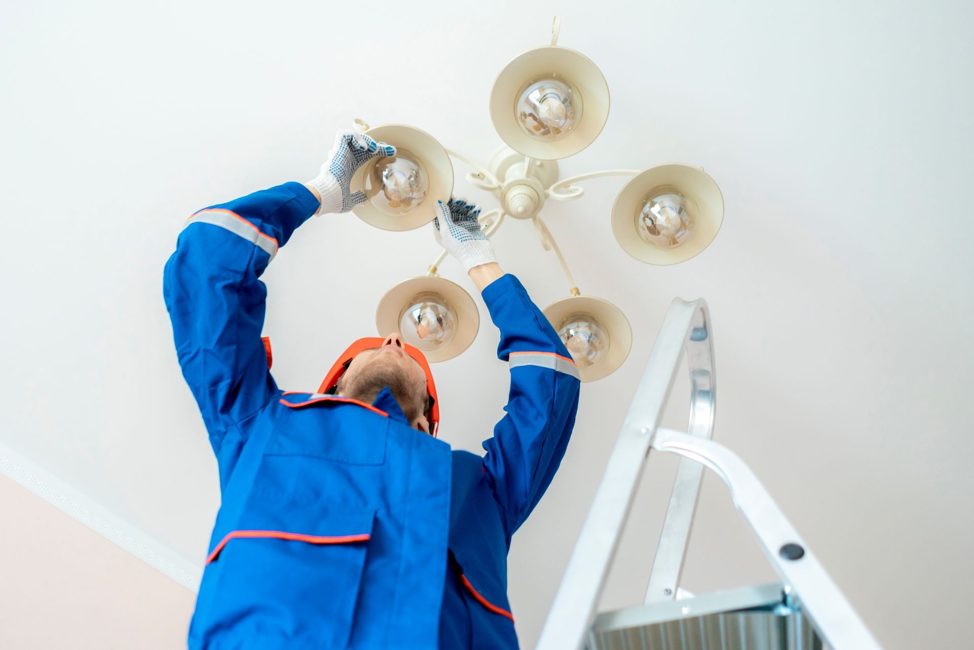 Electrician in blue coveralls replacing a ceiling light fixture on a ladder