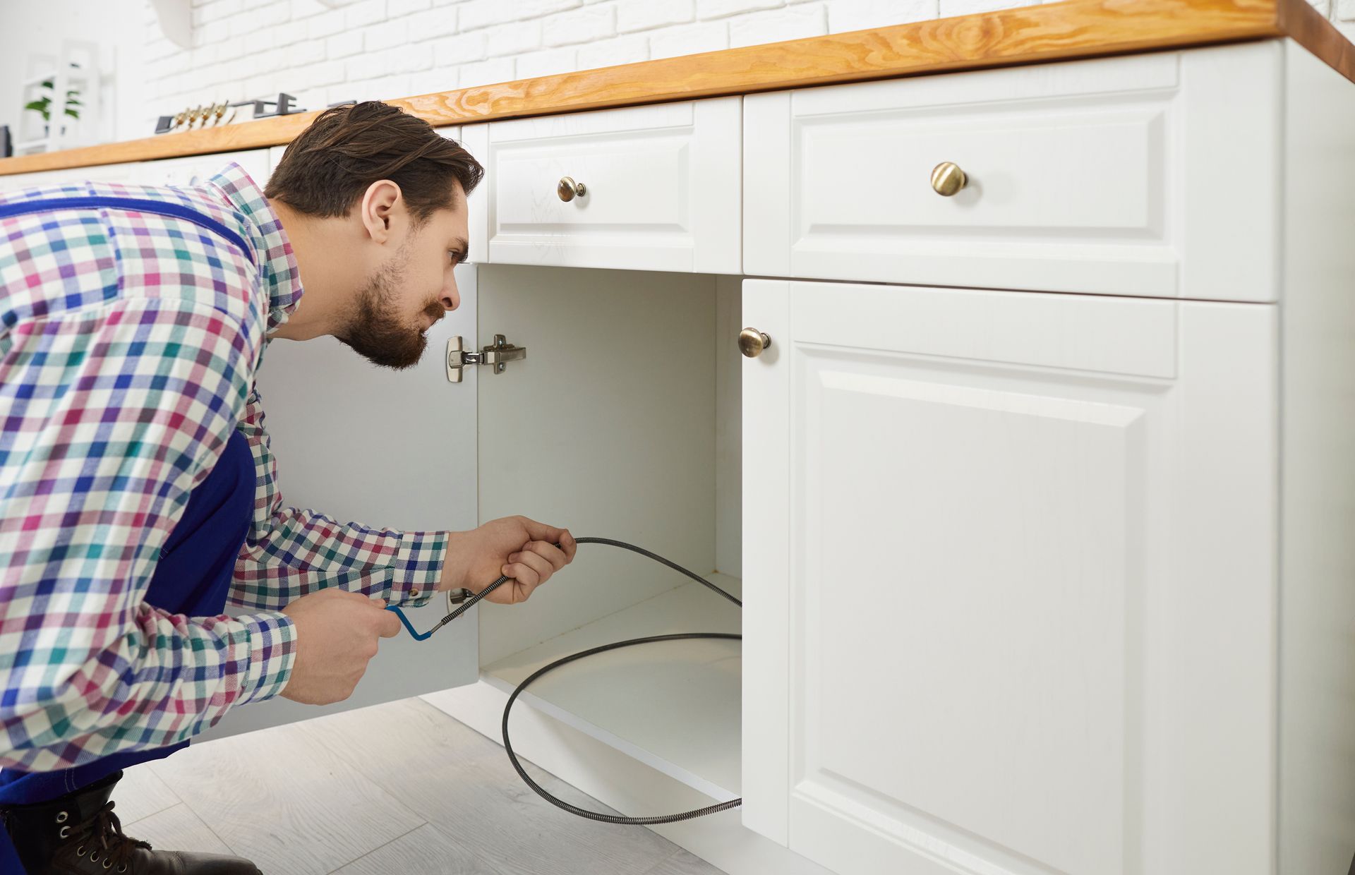 Man crouching under a kitchen cabinet, inspecting wiring with a tool in a white kitchen.
