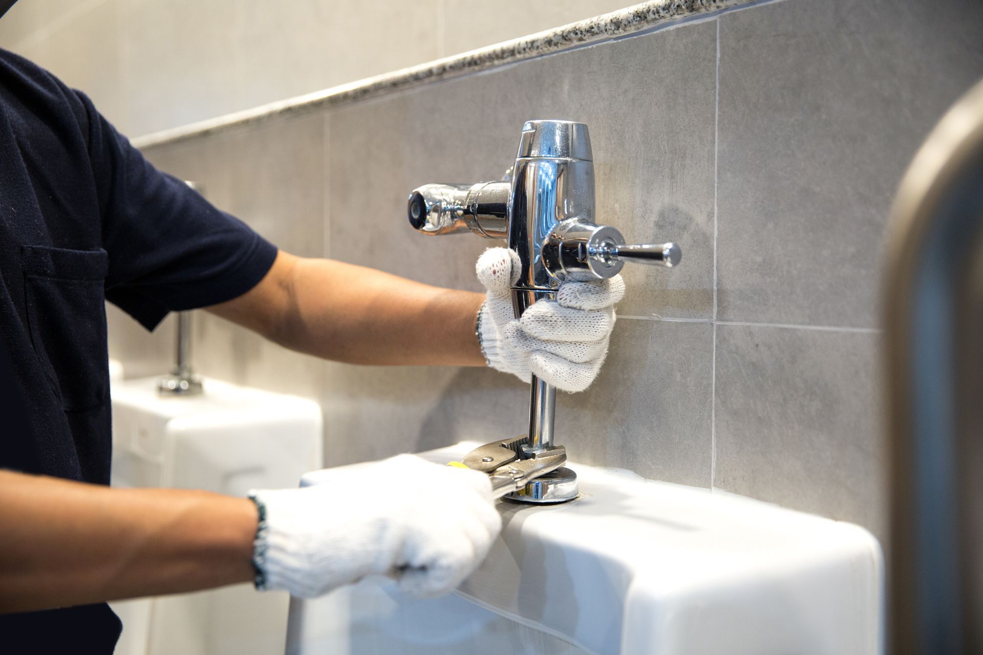Person cleaning a chrome faucet at a white sink with gloves on, in a tiled restroom.