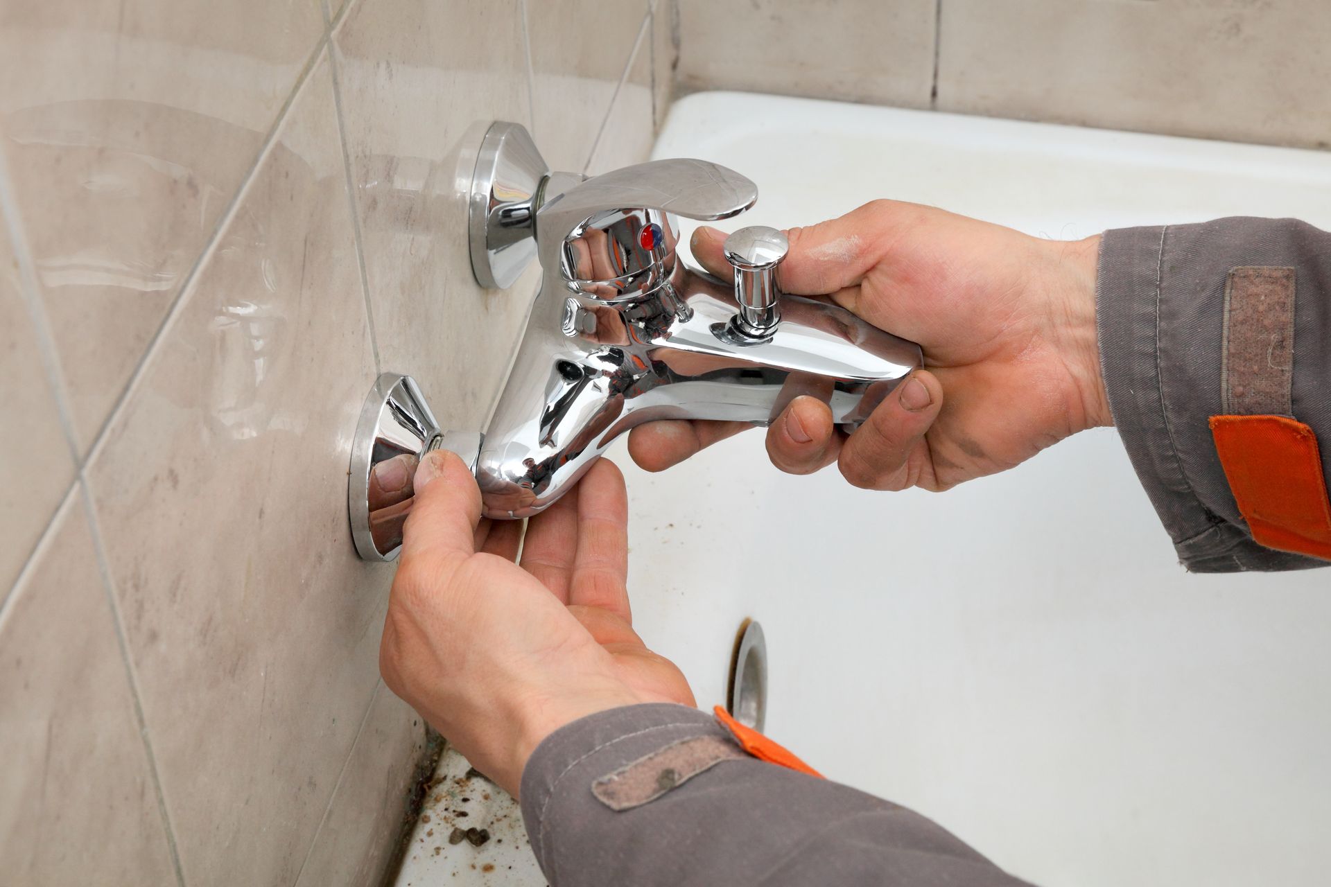 Hands installing a chrome bathtub faucet against beige tile wall