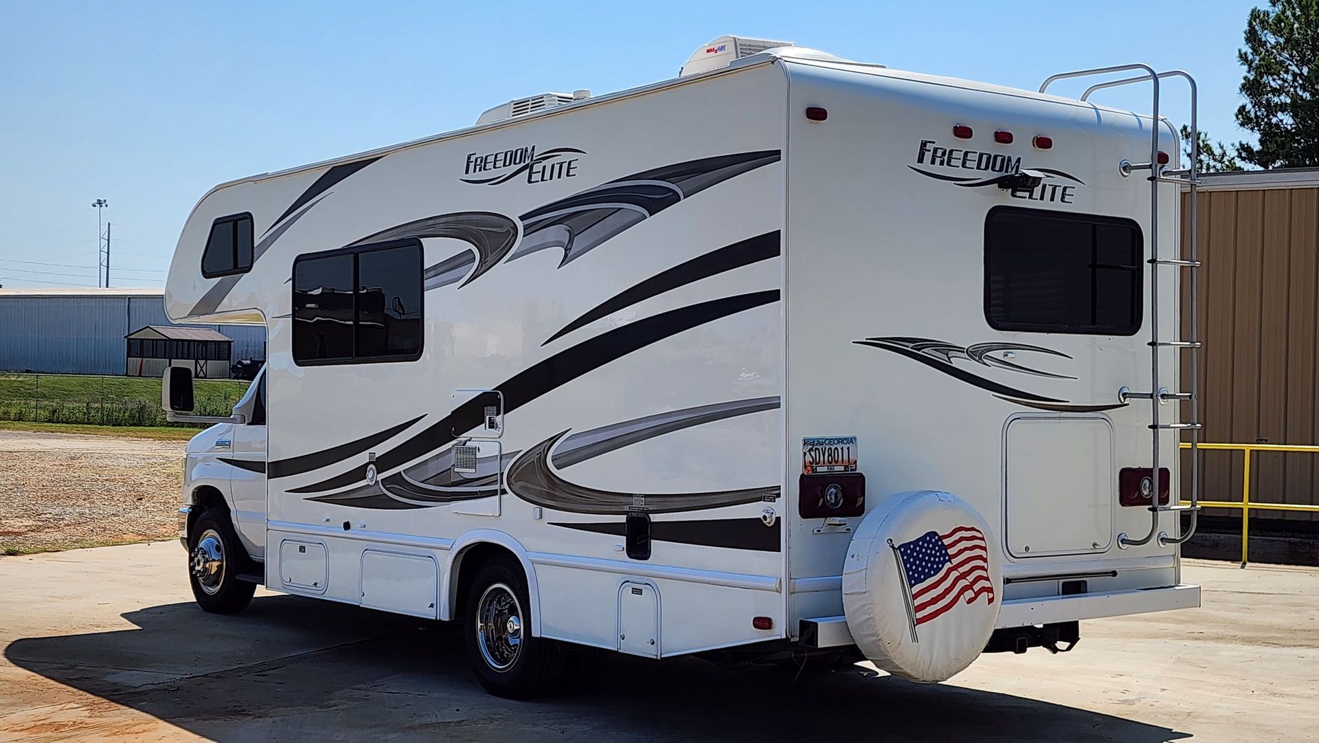White RV with black and gray decals parked on concrete with blue sky. Spare tire cover has American flag.
