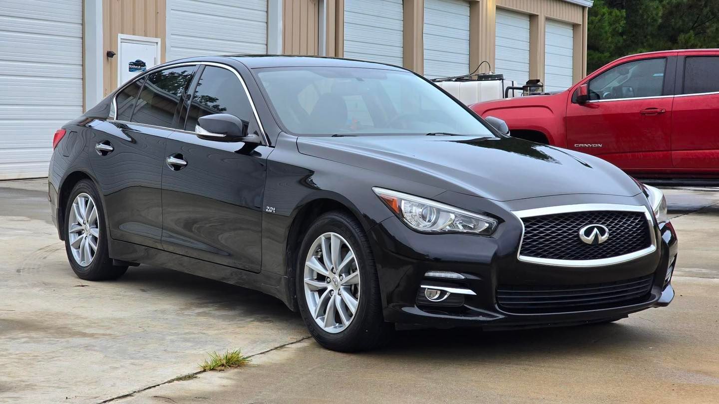 Black Infiniti Q50 sedan parked in front of a tan building with closed garage doors, red truck visible on the right.