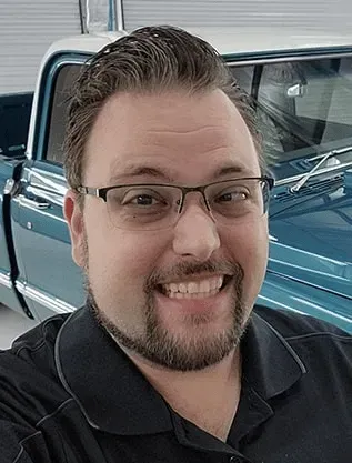 Man with glasses smiles, standing in front of a blue and white classic pickup truck.