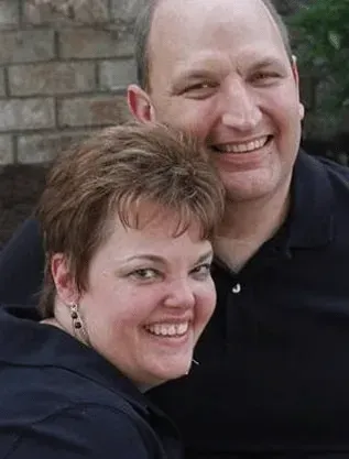 Couple smiling together, woman in front, man behind, brick wall background.