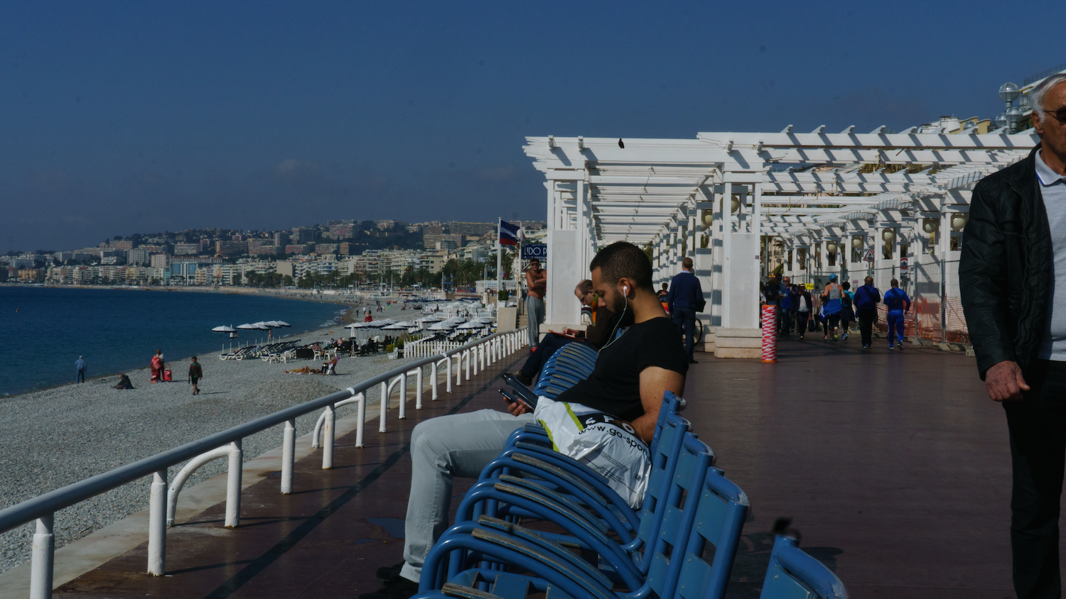 Two people sit on a bench overlooking a beach and city. White pergola in the background. Blue sky.