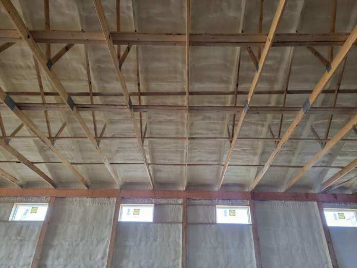 Interior view of a building with exposed wooden beams and spray foam insulation covering the walls and ceiling.