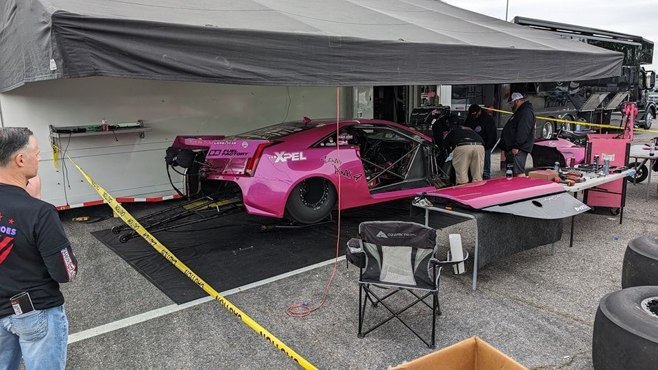 Pink race car being worked on in a pit area, under a canopy. People, tools, and a trailer are visible. | Zoom Automotive