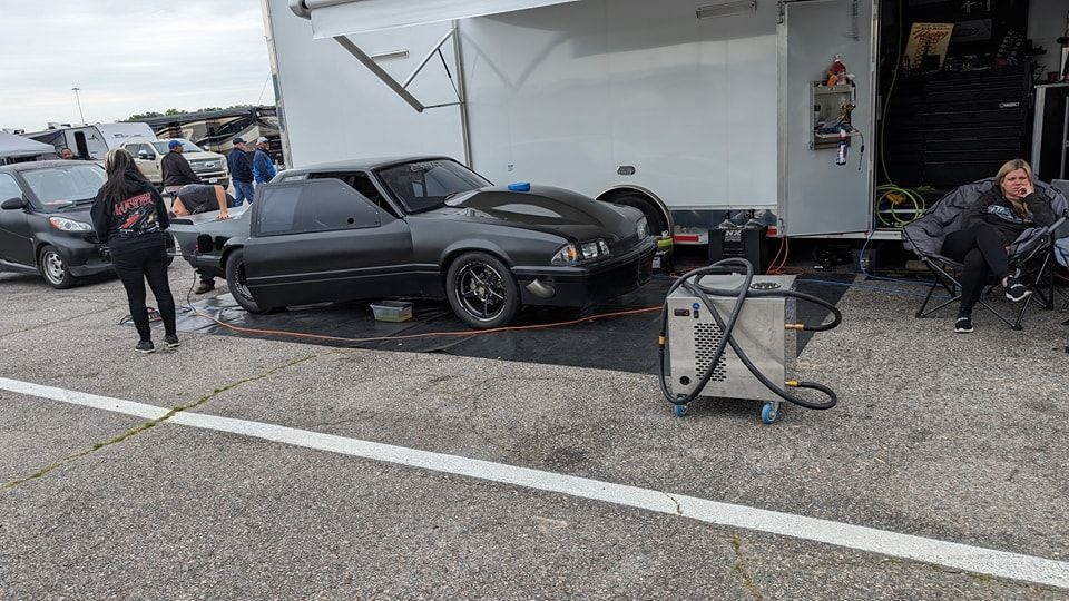 A black drag racing car parked near a trailer at a race track with people nearby. | Zoom Automotive