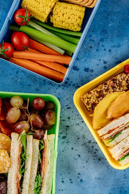 A close up of three lunch boxes filled with sandwiches , fruit , vegetables and crackers.
