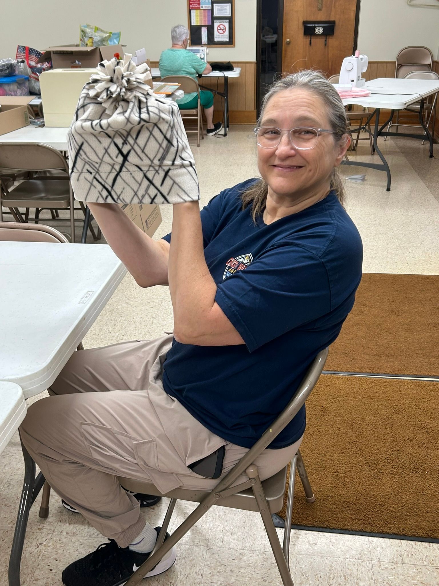 Woman smiles, holding up a handmade cloth item with black and white pattern. She is sitting in a room with sewing equipment.