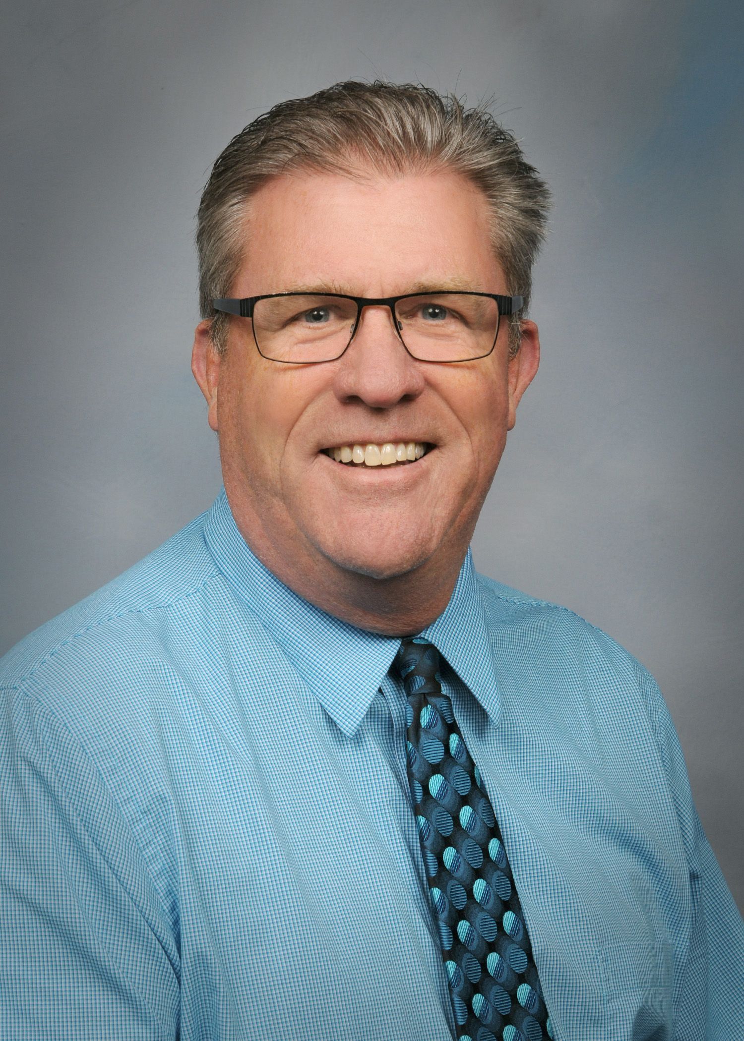 Smiling man in glasses and blue patterned shirt against a gray studio backdrop