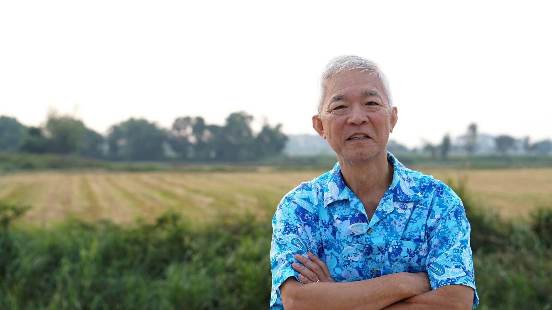 Smiling man in a blue shirt standing with arms crossed in a field outdoors