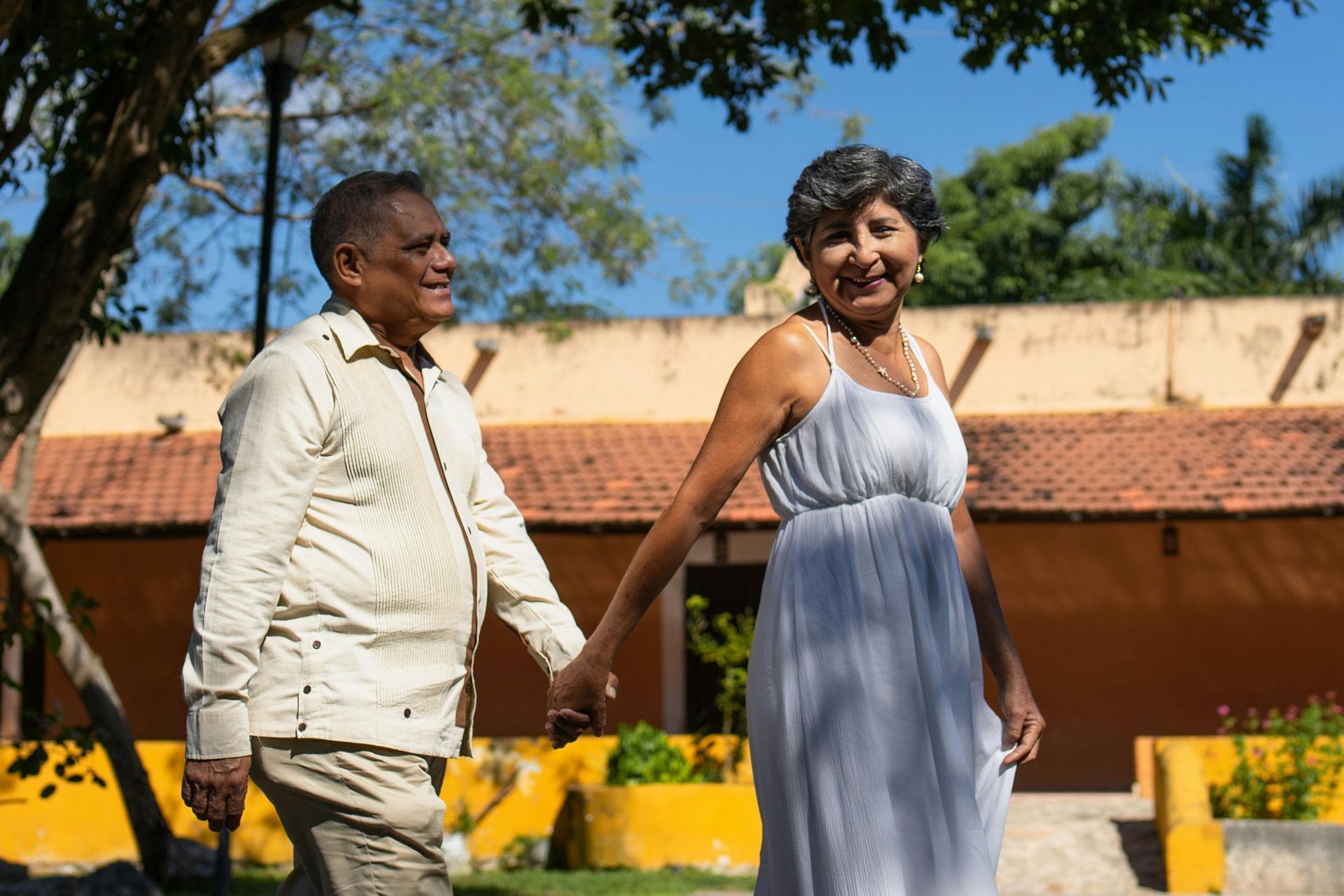 Couple holding hands outdoors by a yellow wall and trees, smiling in sunlight