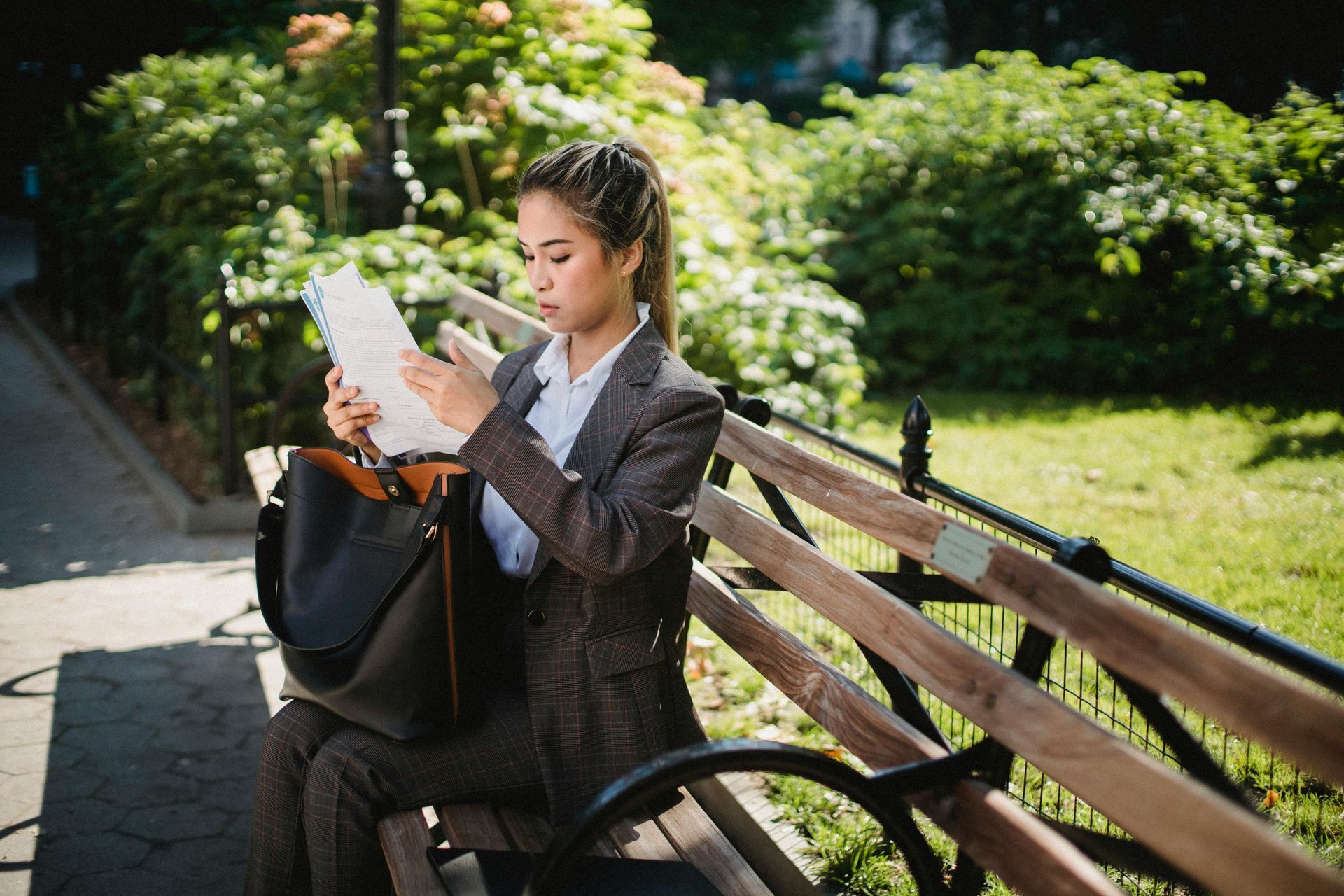 Person reading a map on a park bench beside a black bag and railing in a sunny park