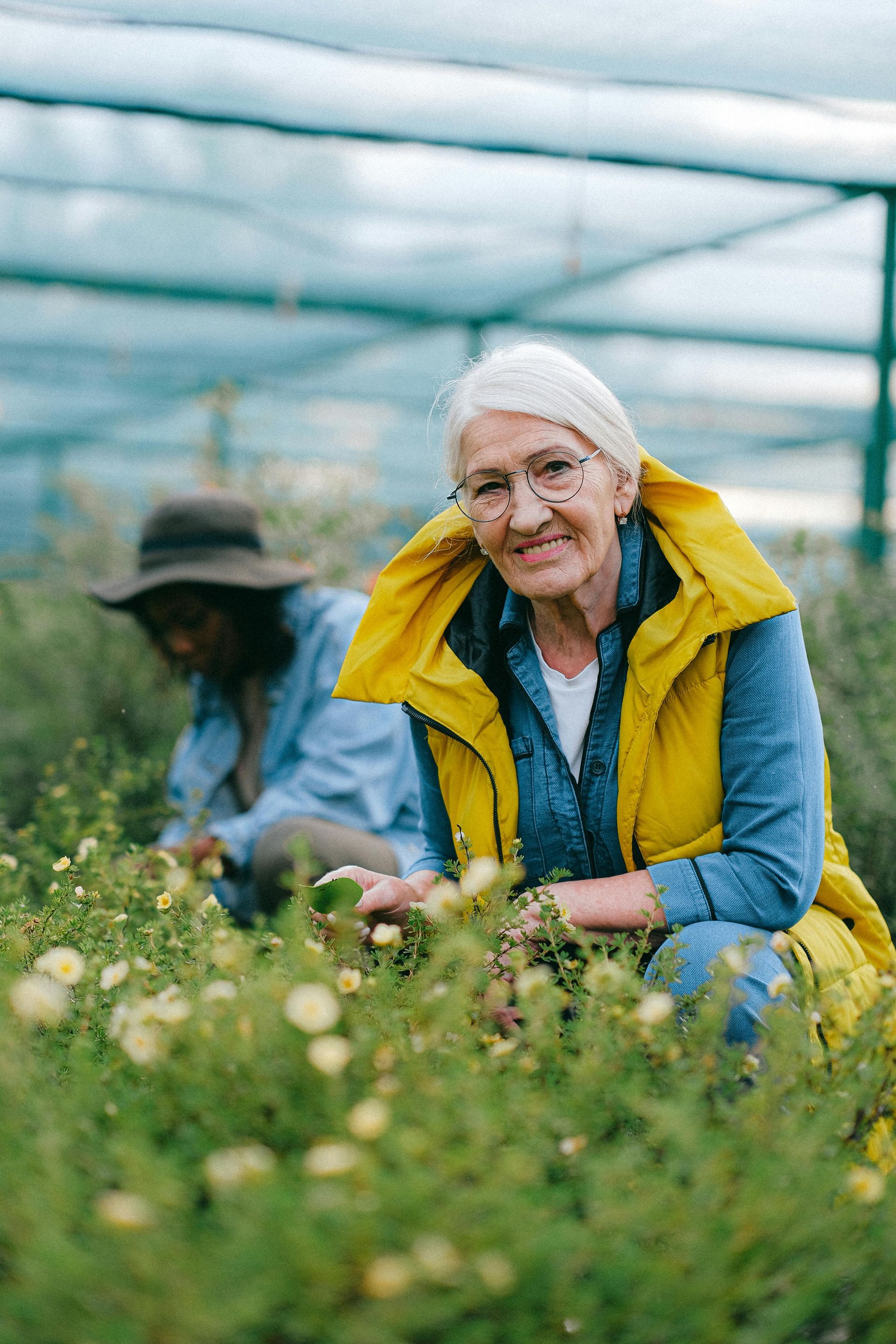 Woman in a yellow jacket kneels among flowers in a greenhouse, smiling at the camera.