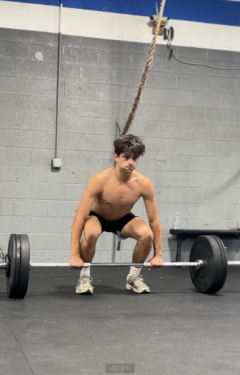 A shirtless man is squatting down to lift a barbell in a gym.