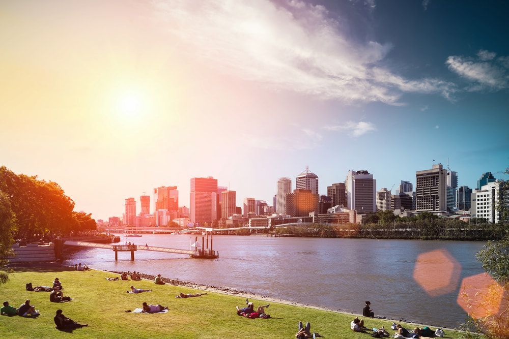 A Group of People Are Sitting on The Grass Near a River with A City Skyline in The Background — Betta Foot Clinic in Sunnybank, QLD 