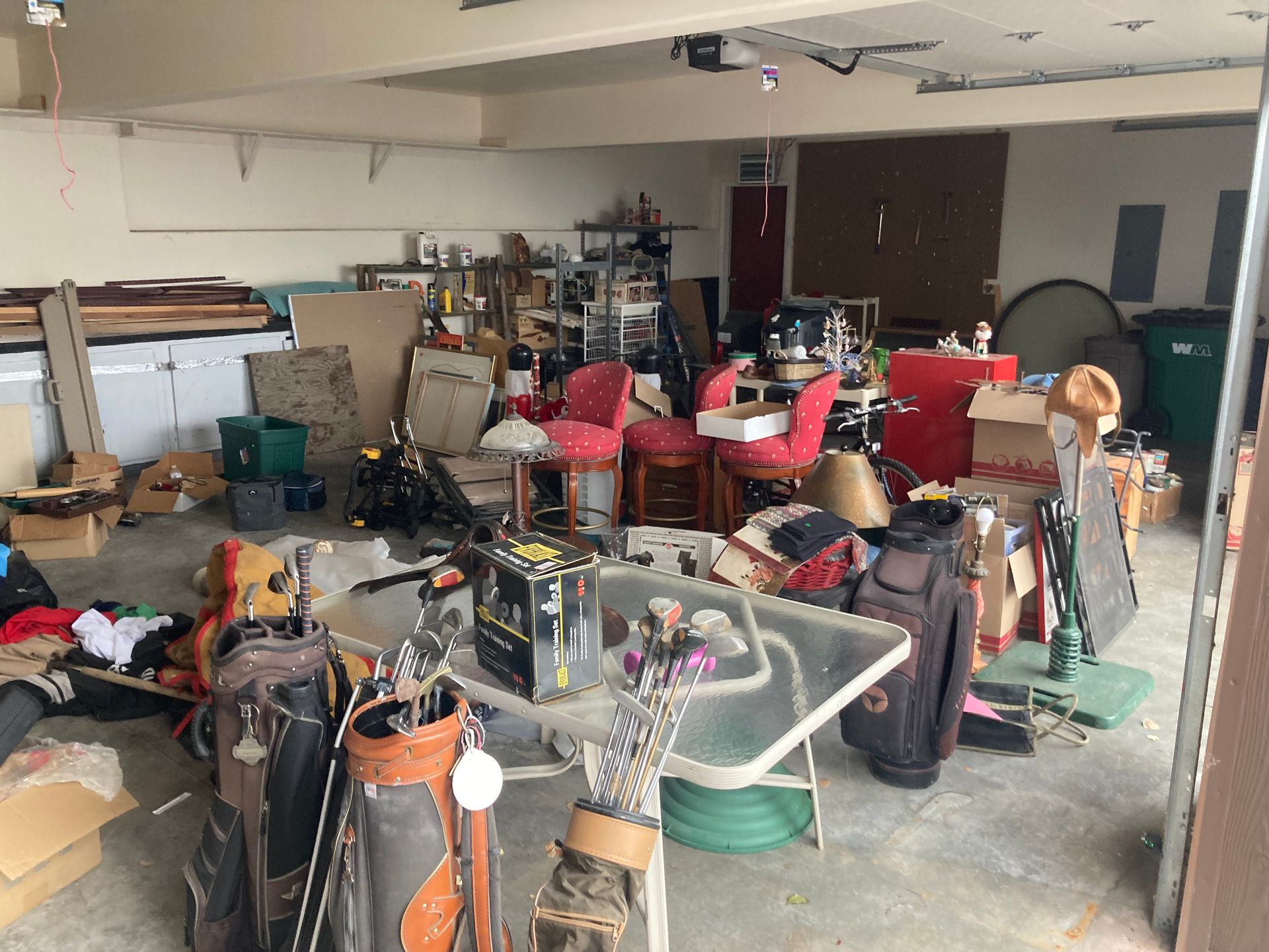 A cluttered garage filled with golf bags, chairs, boxes, and various household items on a glass-topped table and floor.