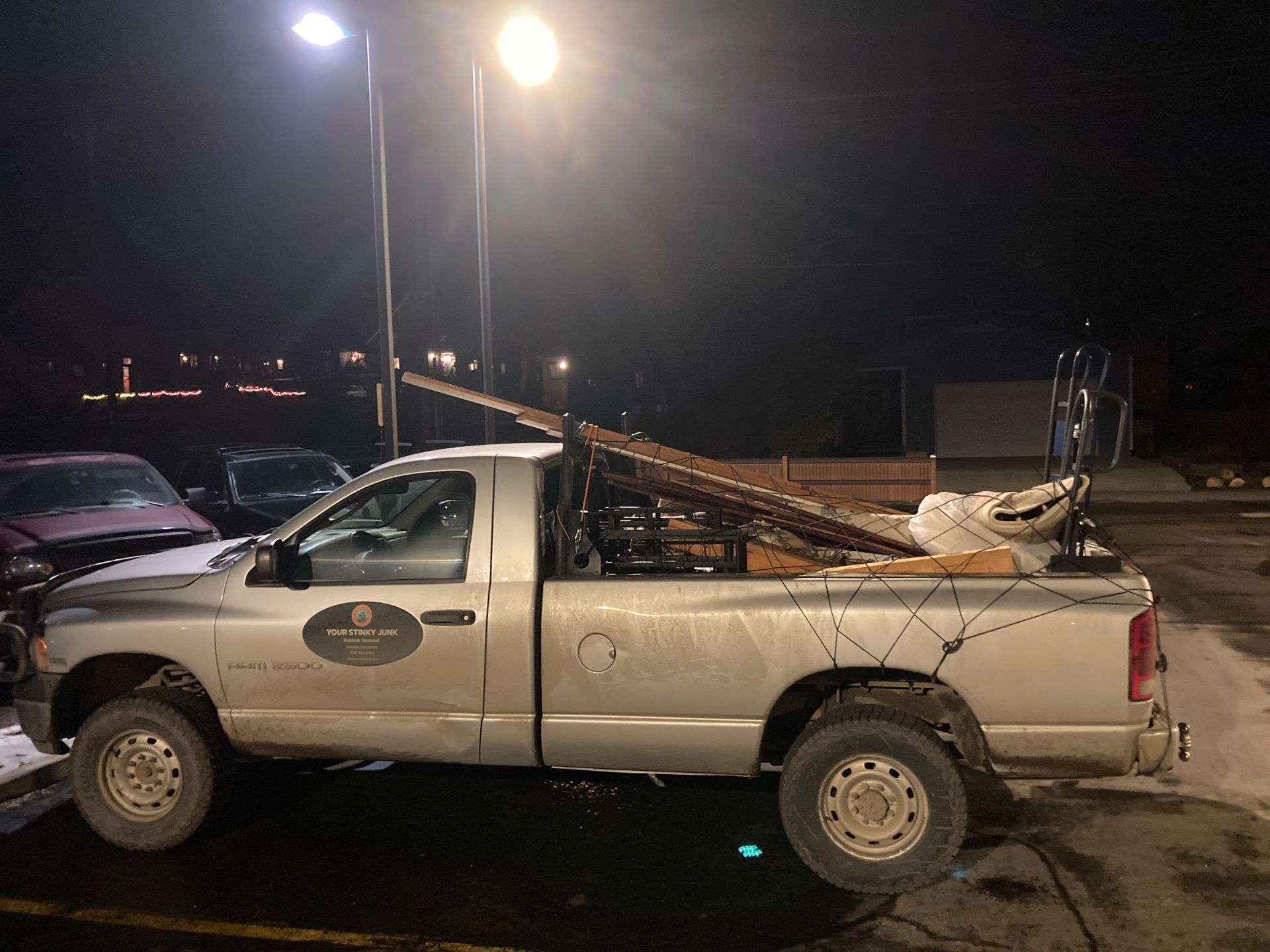 A silver pickup truck parked at night, loaded with a long ladder, miscellaneous equipment, and cobweb decorations.