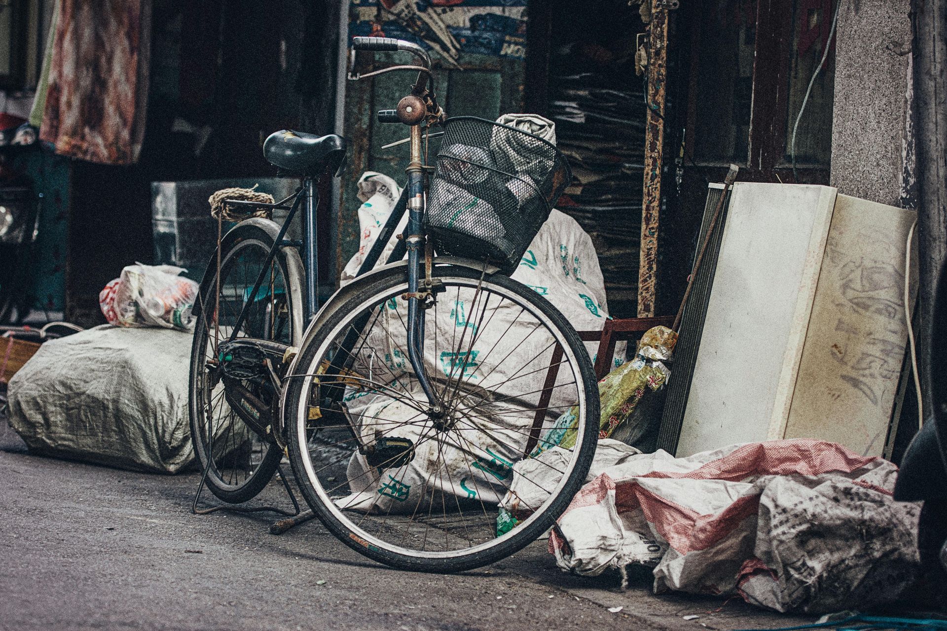 A black bicycle with a front basket parked on a street next to several large, cluttered bags of discarded items.