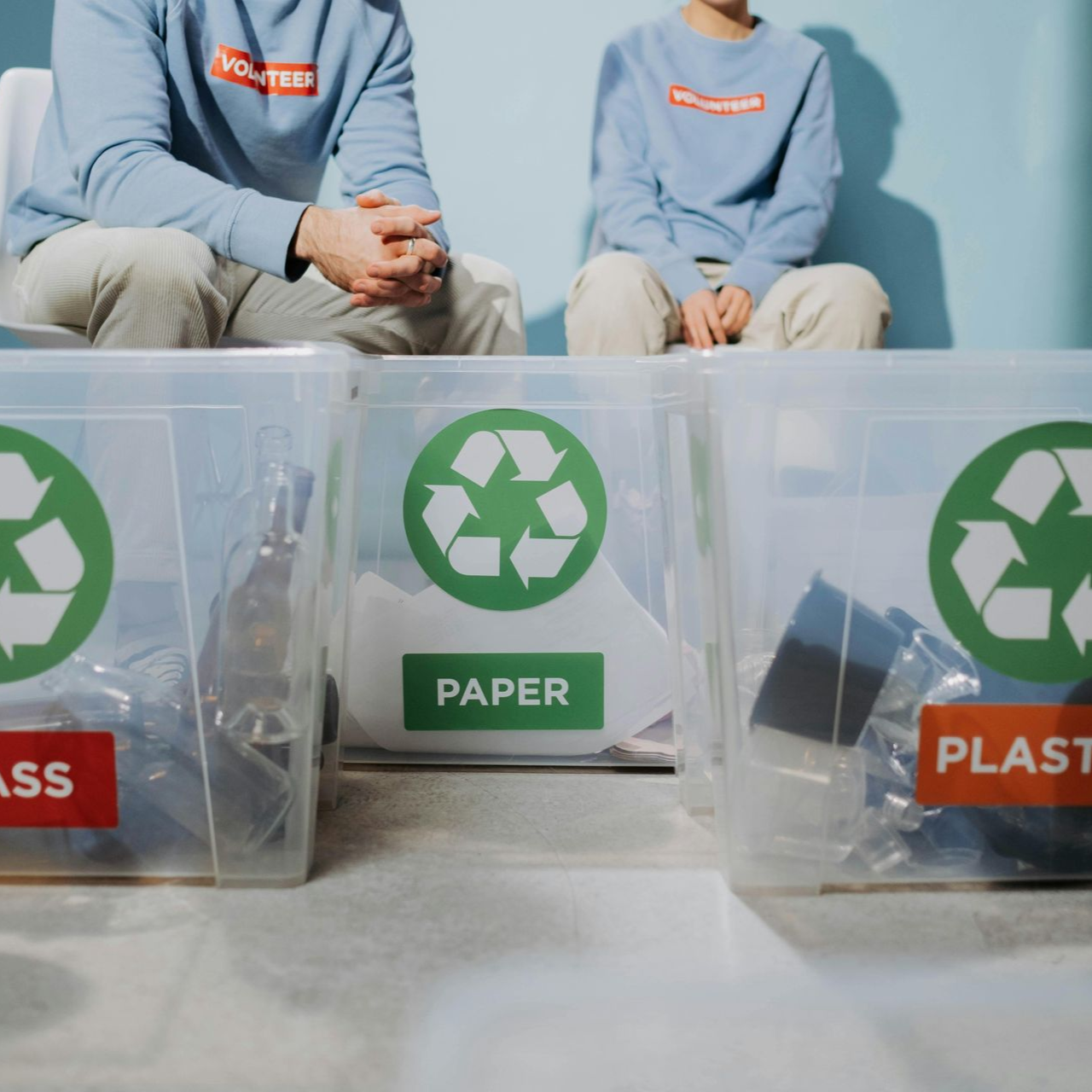 Two volunteers sit behind three clear recycling bins labeled for glass, paper, and plastic.