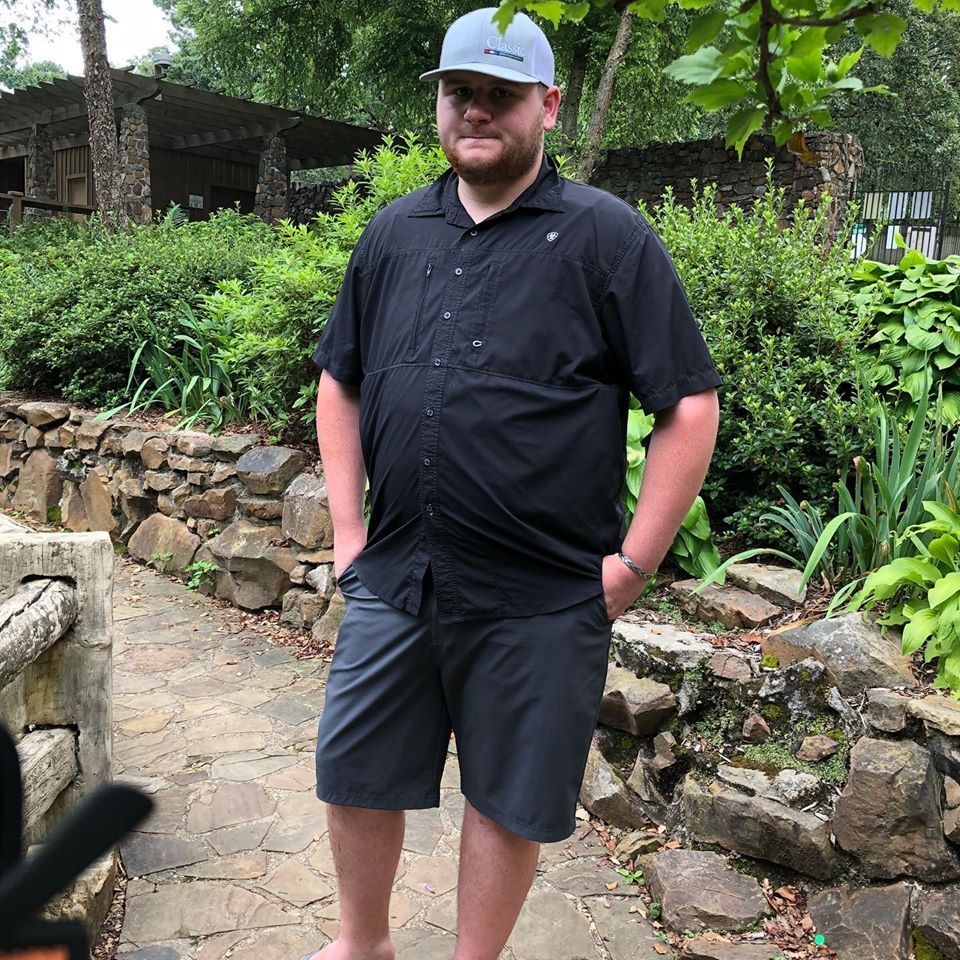 A man in a black shirt and shorts is standing in a garden with his hands in his pockets.