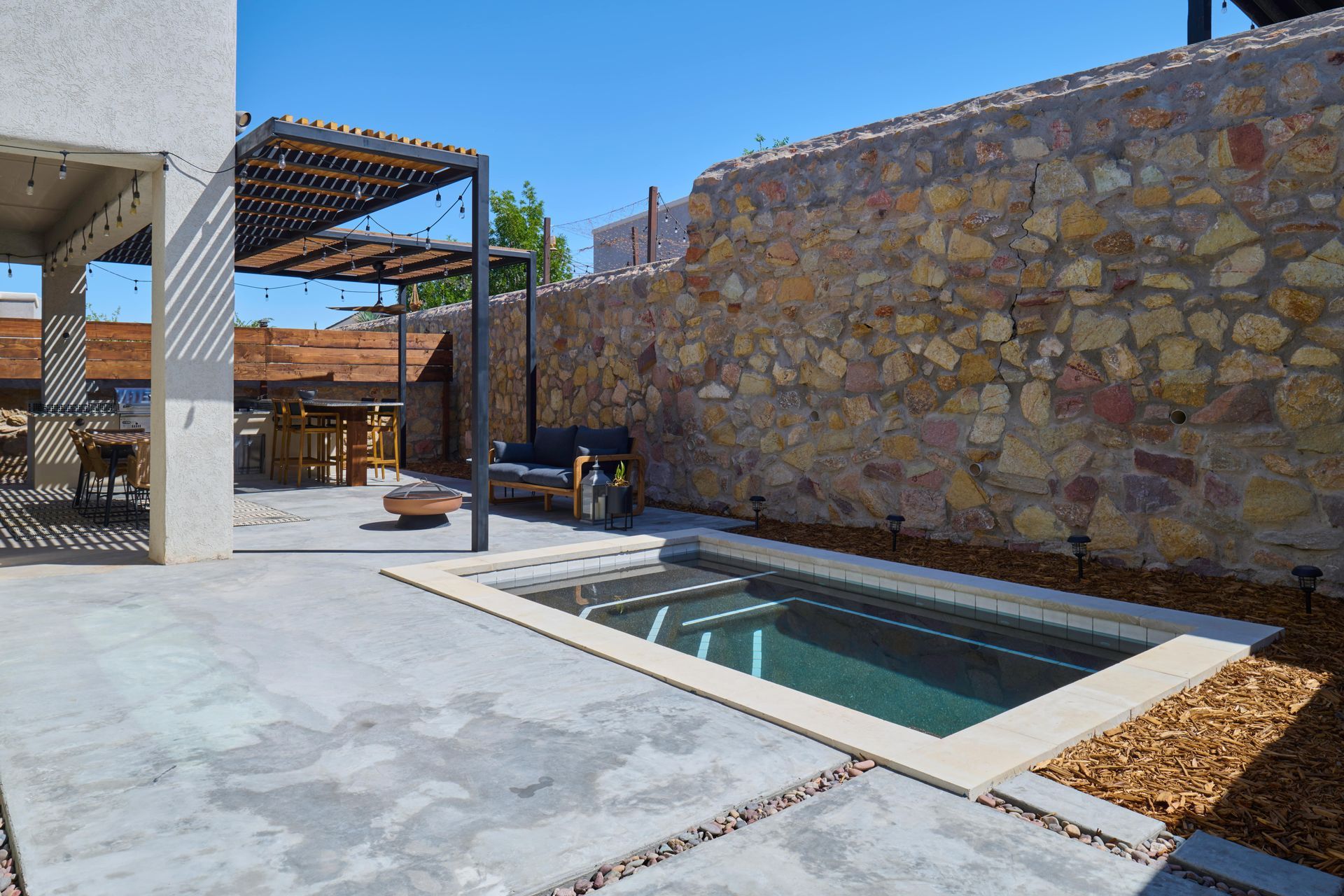 Backyard with small pool, patio, stone wall, and pergola under a clear blue sky.