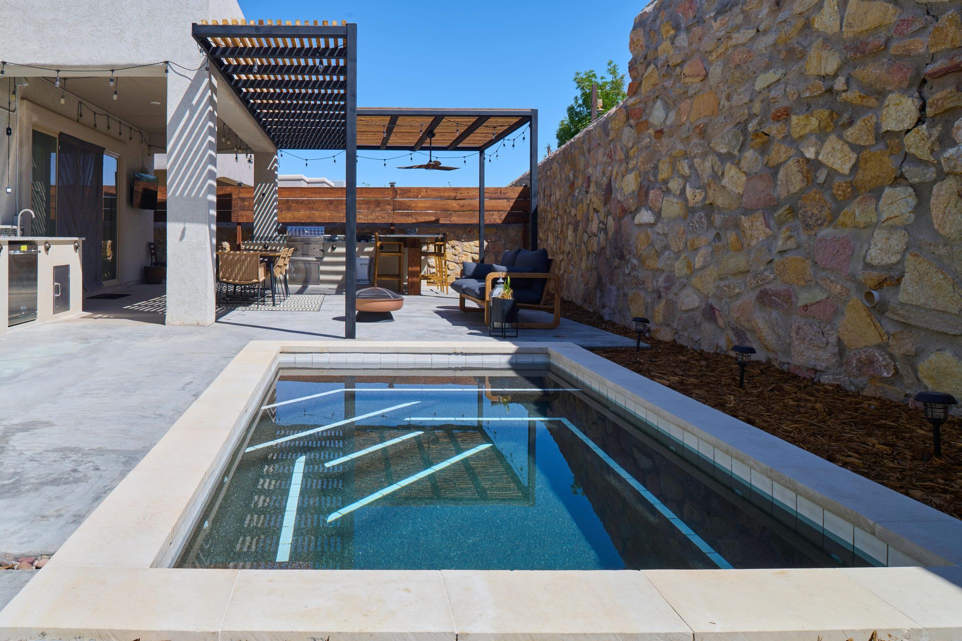 Backyard with rectangular pool, patio, and stone wall. Pergola over seating area.