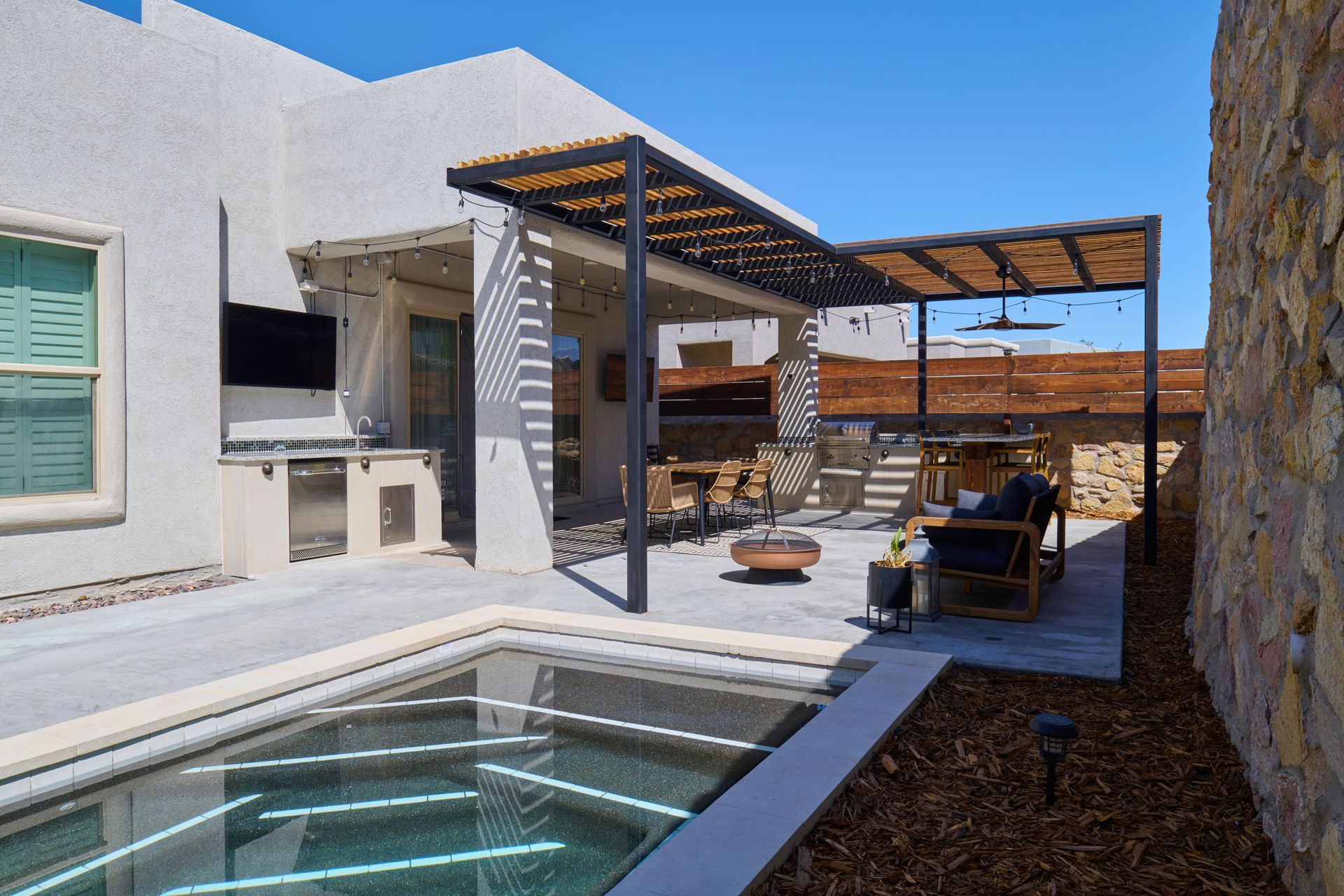 Outdoor patio with pool, pergola, seating, and built-in kitchen area. Bright sunlight and blue sky.