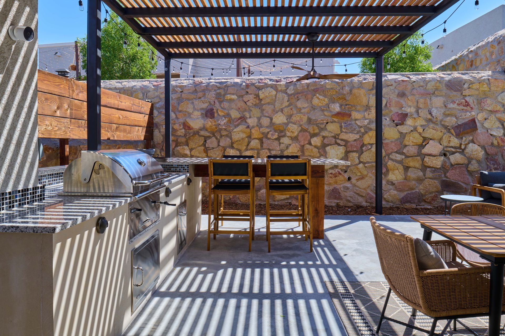 Outdoor kitchen with grill, bar, and stone wall, beneath a pergola.