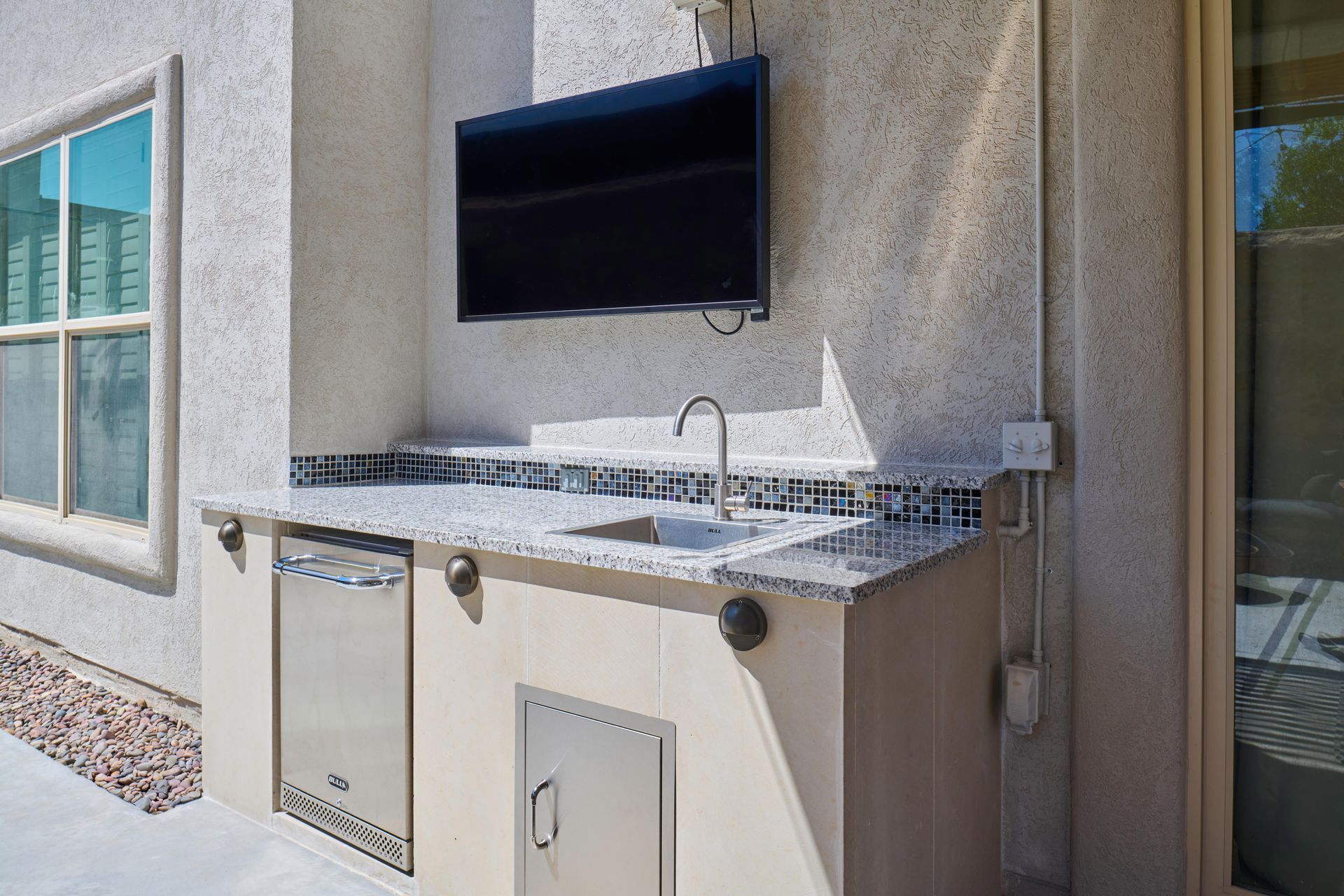 Outdoor kitchen with TV, sink, cabinets, and appliances on a stucco wall.