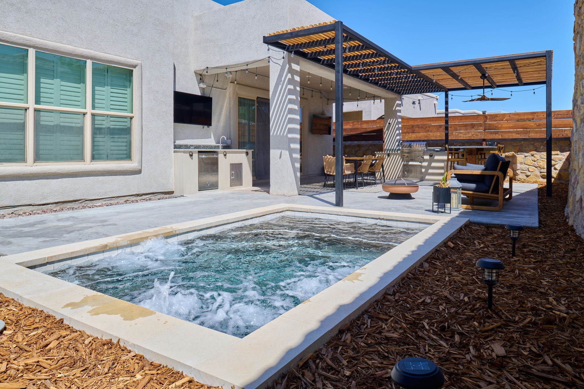 Outdoor jacuzzi with pergola, patio furniture, and landscaping against a stucco home.