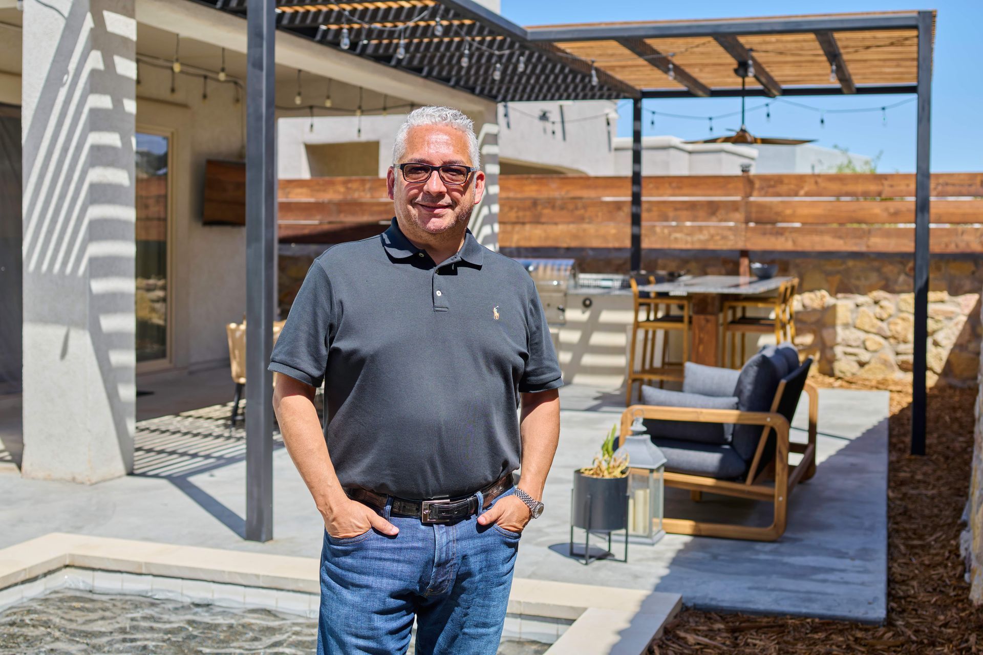 Man standing by a pool in an outdoor setting, smiling, arms at sides, gray shirt, and jeans.