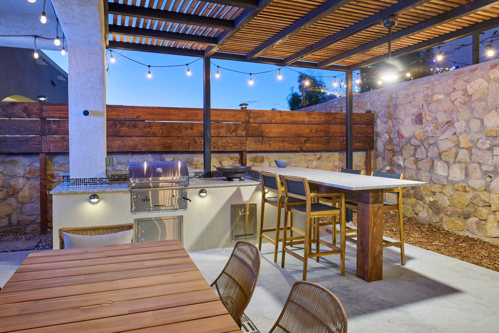 Outdoor kitchen with grill, dining table, and string lights under a pergola.