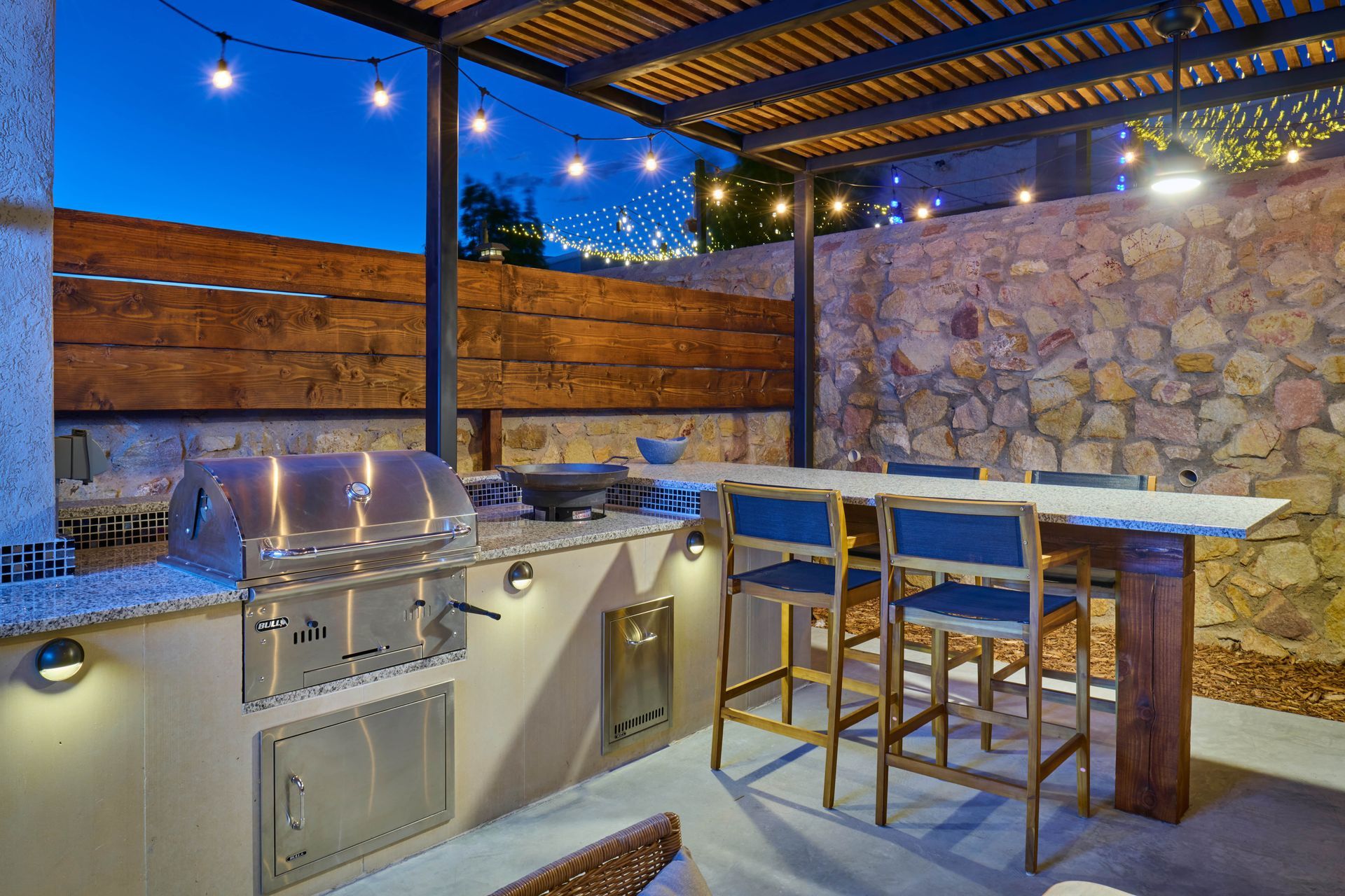 Outdoor kitchen with grill, counter, and seating under a pergola, lit by string lights at dusk.