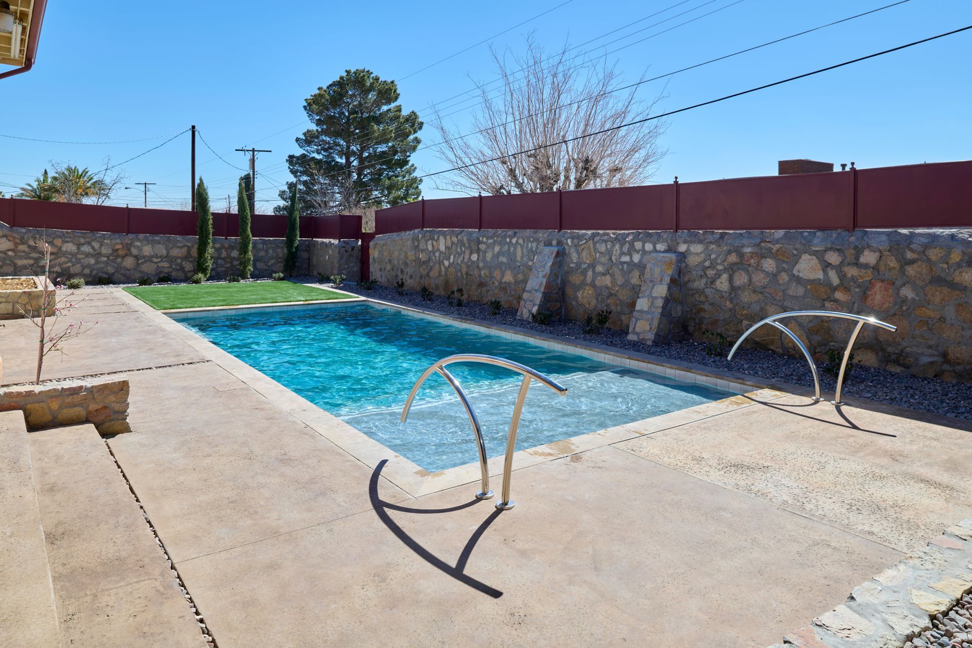 A bright blue rectangular swimming pool with metal handrails sits on a concrete patio bordered by a stone wall.
