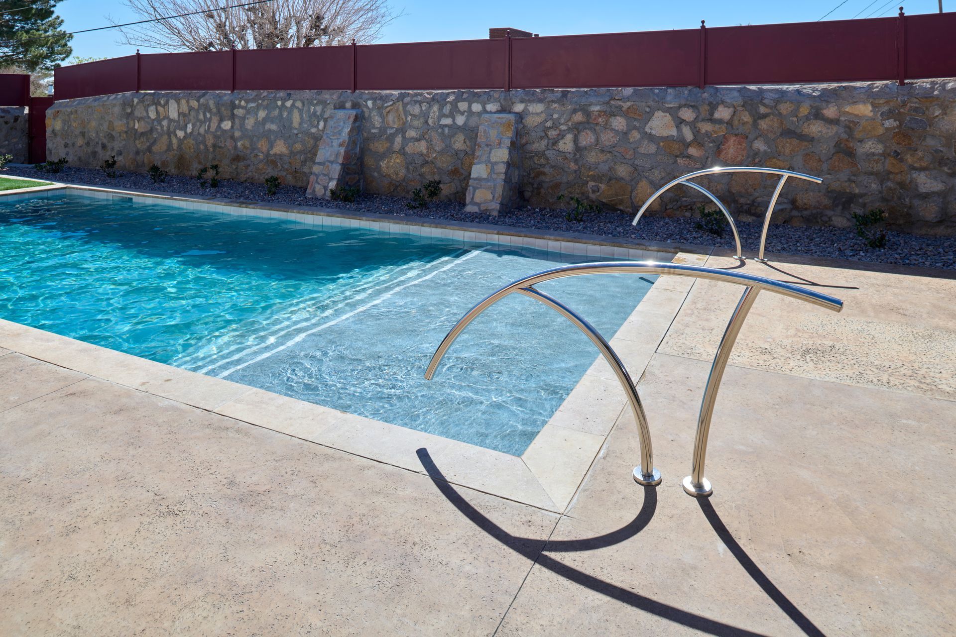 A turquoise swimming pool with a Baja shelf, featuring modern curved metal handrails on a concrete deck.
