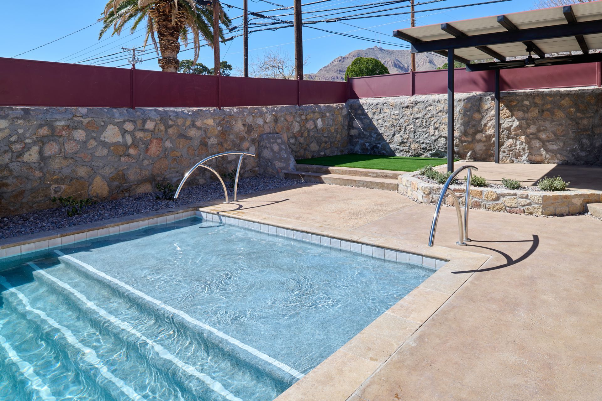 A stone-walled pool area with light blue water, steps, metal handrails, and a shaded patio under a clear blue sky.