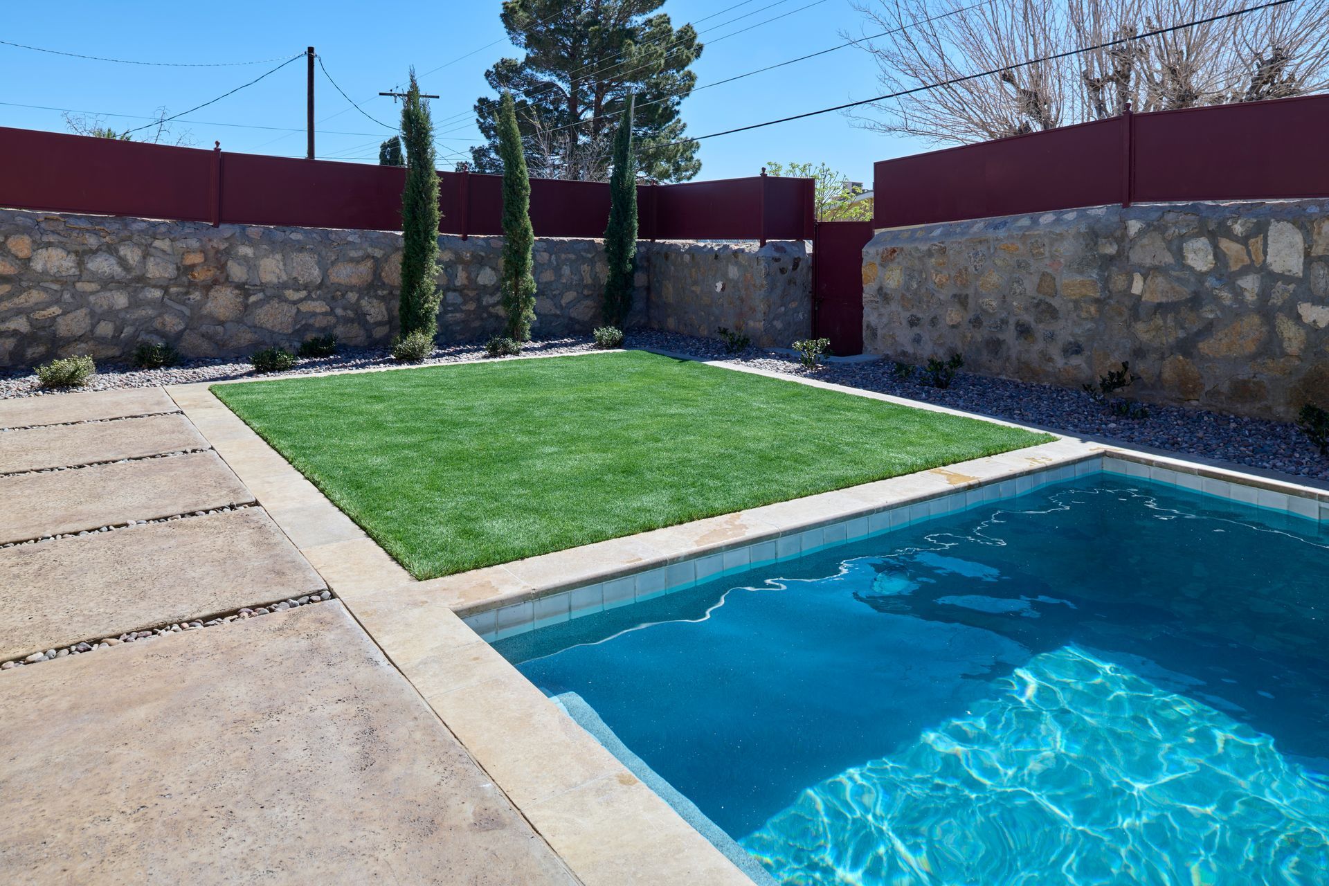 A bright blue swimming pool sits next to a rectangular patch of green grass, bordered by stone walls and a reddish fence.
