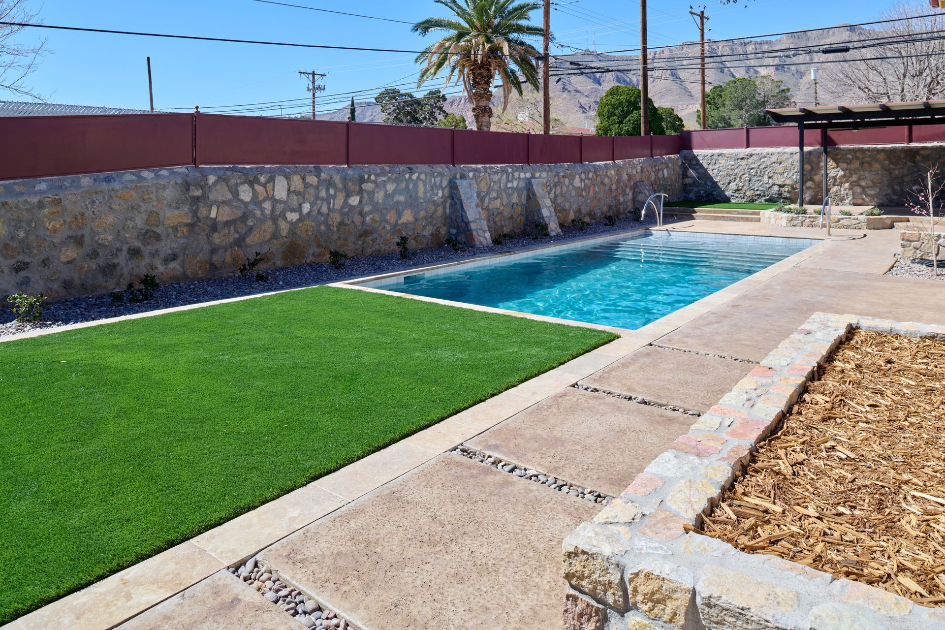 A rectangular swimming pool surrounded by a stone wall, green turf, and paved patio area under a bright, sunny sky.