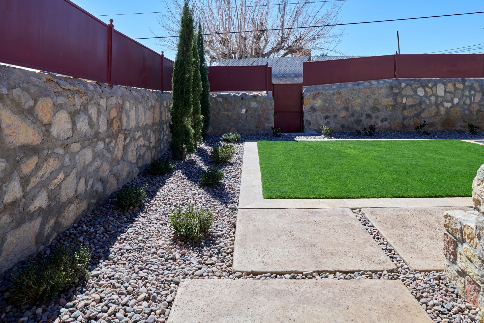 A backyard with a stone wall, red fencing, a patch of green grass, concrete pavers, and gravel landscaping.