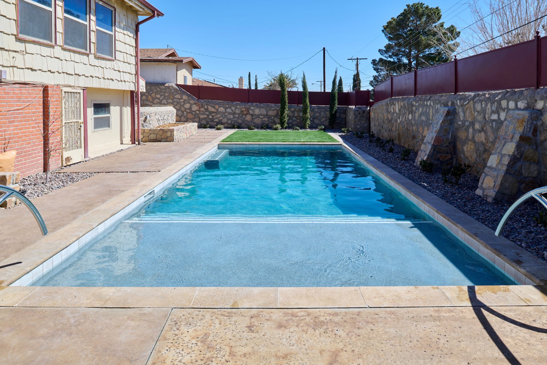 Rectangular swimming pool with a shallow sun shelf, set in a backyard with stone retaining walls and a brick house.