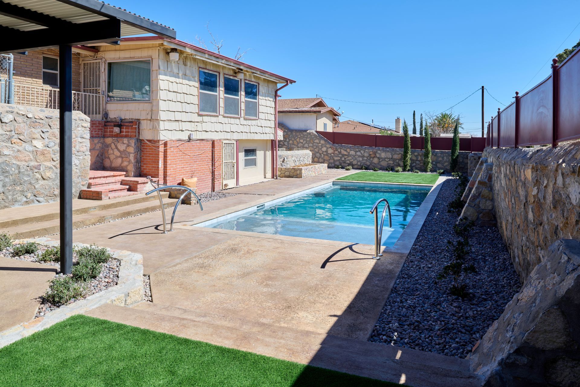 A sunny outdoor backyard with a rectangular swimming pool, stone retaining walls, and a multi-story house.
